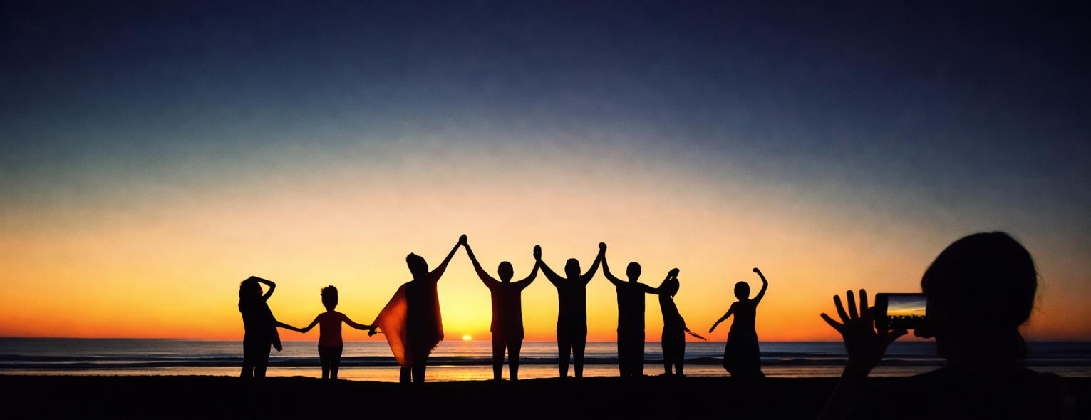Silhouettes on a beach at sunset: a group in the distance and someone in the foreground taking a photo on a phone—Soil and Sky, less screen time, more nature.